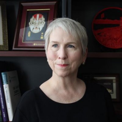 woman with short hair looks off camera, there is a bookshelf and framed images behind her